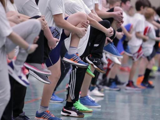 Schülerinnen und Schüler stehen beim Sportunterricht in einer Reihe, alle auf einem Bein stehend.