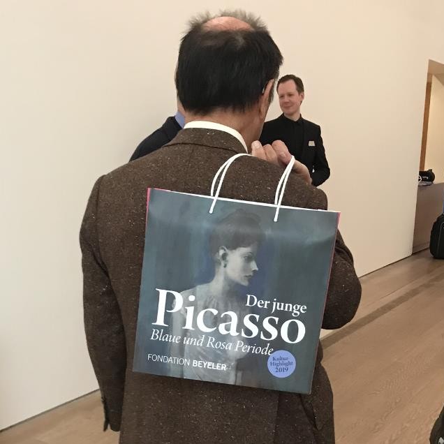 Claude Picasso, Sohn von Pablo Picasso, hält eine Tragetasche in der Hand, die mit Titel und Titelmotiv der Ausstellung bedruckt ist.