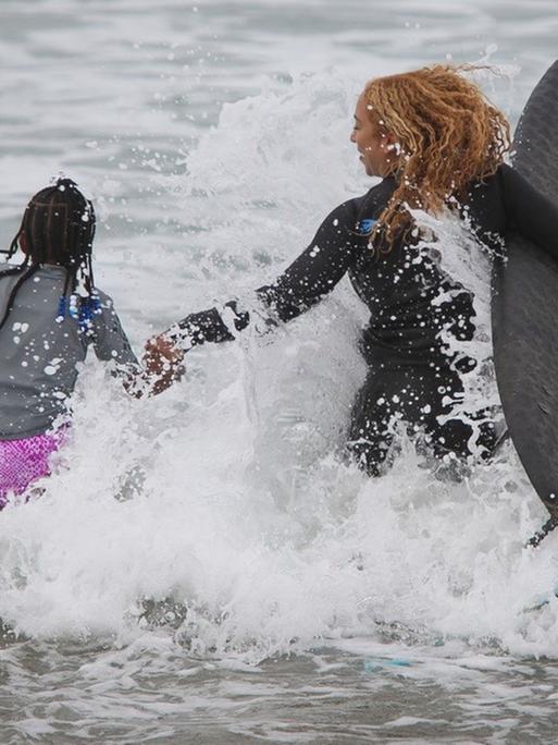 Die Surflehrerin und ihre Schülerin laufen in das spritzende Wasser.