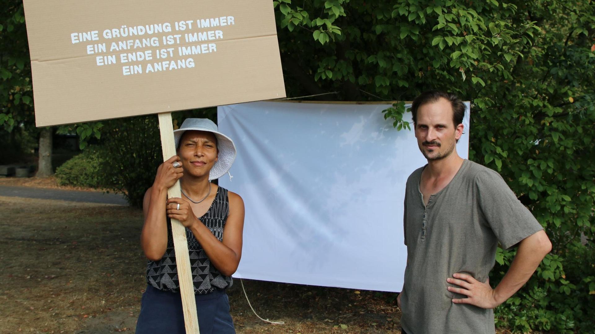 Artists in Residence Joyce Sadiq und Sebastian Bodirsky halten ein Schild mit der Aufschrift: "Eine Gründung ist immer ein Anfang"