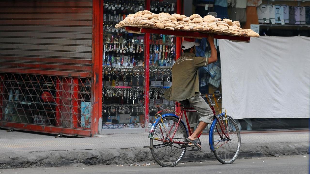 Ein Brotverkäufer balanciert ein großes Brettgestellt voll beladen mit Brot auf seinem Kopf und fährt mit dem Fahrrad durch eine Straße in Kairo zur Kundschaft.