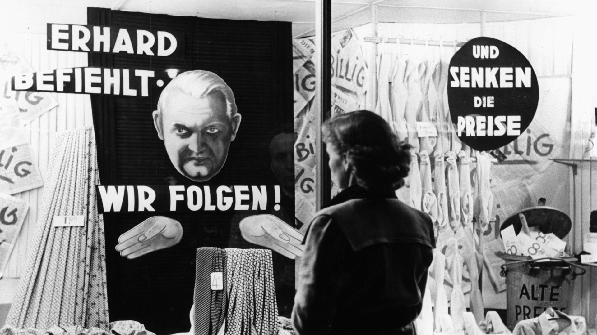 Eine Frau steht vor einem Schaufenster. In dem Schaufenster steht "Erhard befiehlt - wir folgen! und senken die Preise". Das Foto ist von circa 1949/50.