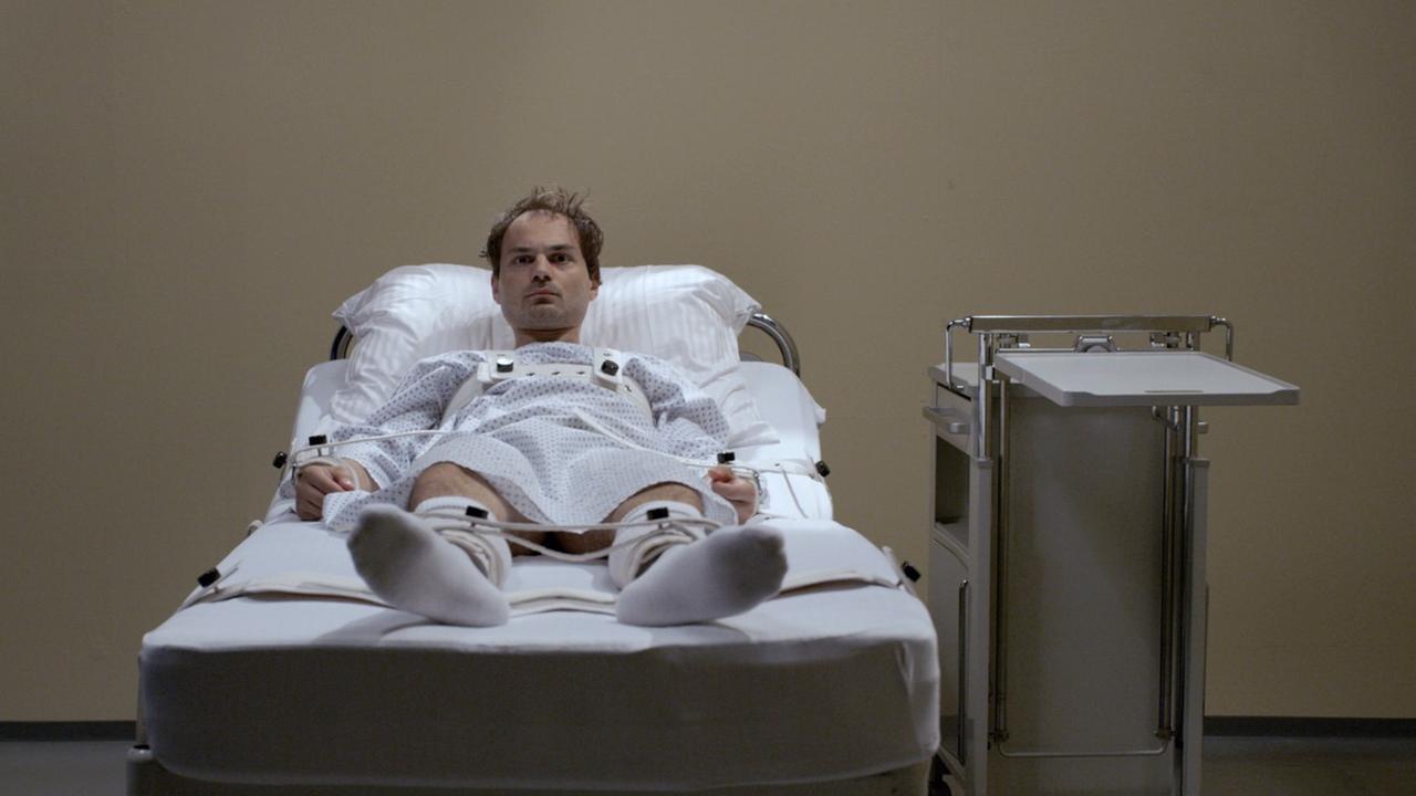 Die Szene aus Rosa von Praunheims Film Darkroom zeigt einen Mann in einem Krankenbett liegend und frontal in die Kamera schauend.