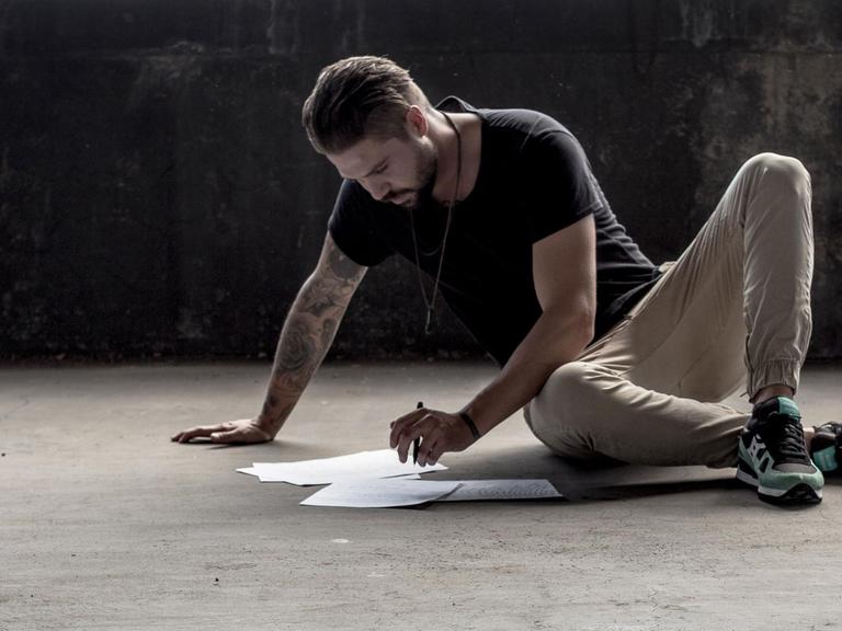 Ein Mann sitzt auf einem Betonboden und schreibt auf einigen Blättern, die er vor sich ausgebreitet hat.