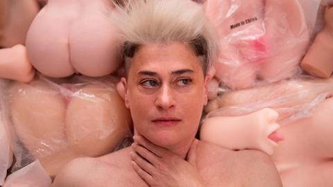 Auf dem Bild ist die Musikerin Peaches zu sehen. Hinter ihr befinden sich viele Sexpuppen.