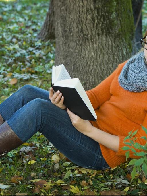 Junge Frau mit Brille sitzt im Park und liest ein Buch.