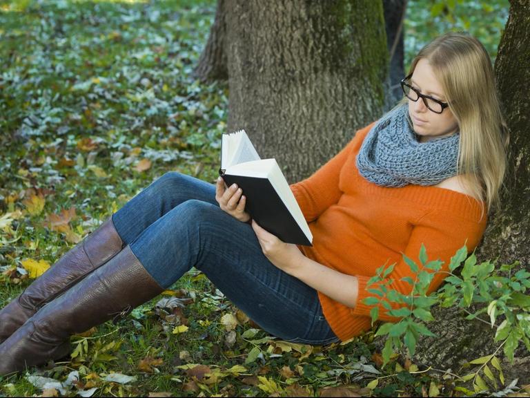 Junge Frau mit Brille sitzt im Park und liest ein Buch.