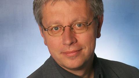 Ein Mann mit grauen Haaren und Brille steht in grauem Anzug vor himmelblauem Hintergrund.