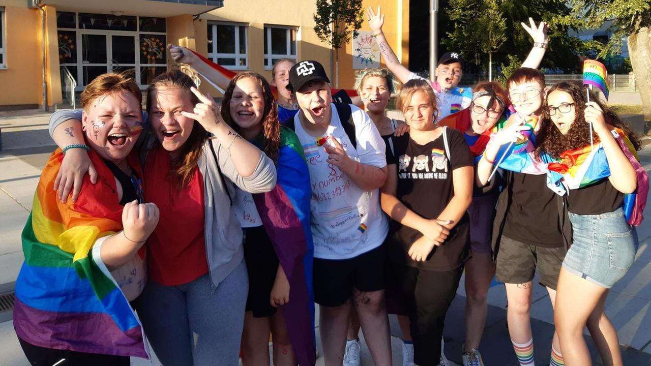 Eine Gruppe junger Menschen mit Regenbogenfahnen vor einer Schule