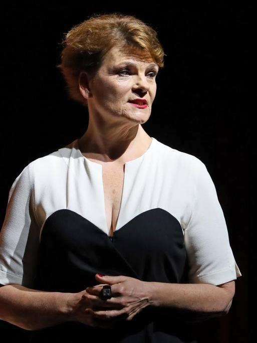 Eine mittelalte Frau in schwarz-weißem Oberteil und mit Kurzhaarschnitt auf einer Bühne schaut ernst zur Seite.