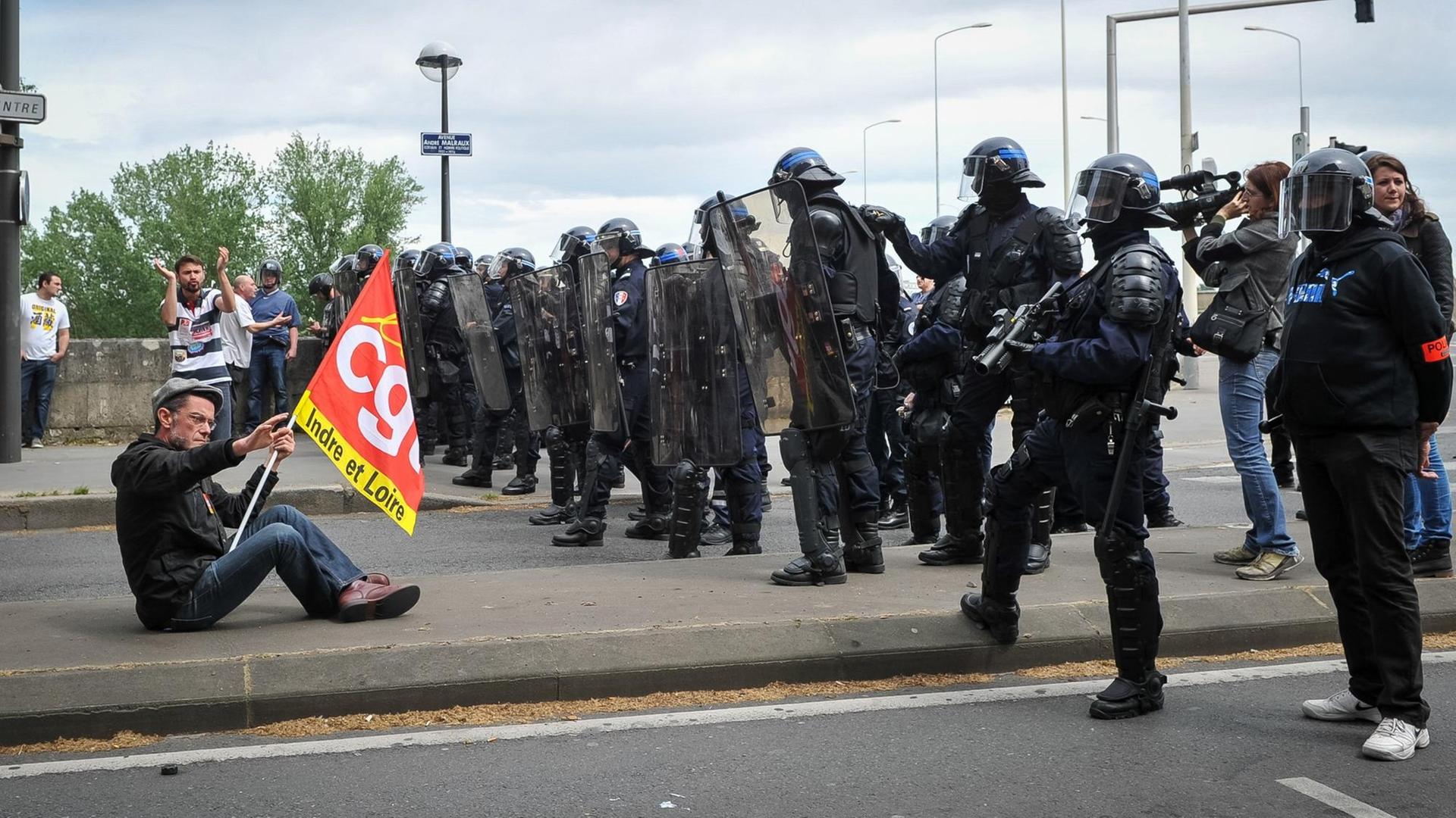 Ein Demonstrant sitzt mit einer Flagge auf einer Straße, vor ihm eine Reihe Polizisten.