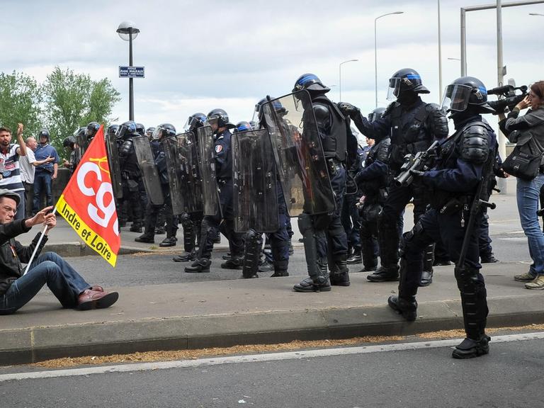 Ein Demonstrant sitzt mit einer Flagge auf einer Straße, vor ihm eine Reihe Polizisten.