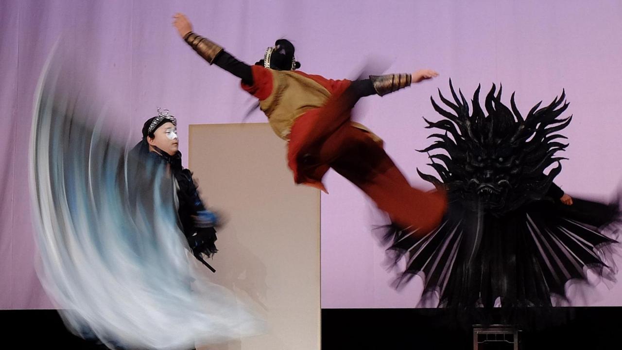 Über die Bühne in der Bildmitte springt ein rot kostümierter Darsteller mit ausgebreiteten Armen durch die Luft, links schwingt ein anderer Darsteller ein verschwommmenes blaues Tuch 