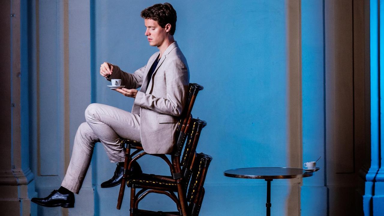 Ein Mann sitzt auf dem obersten von drei übereinander gestapelten Stühlen und hält eine Teetasse vor sich. Man sieht ihn seitlich, der Hintergrund ist bläulich, er trägt einen hellen Anzug.