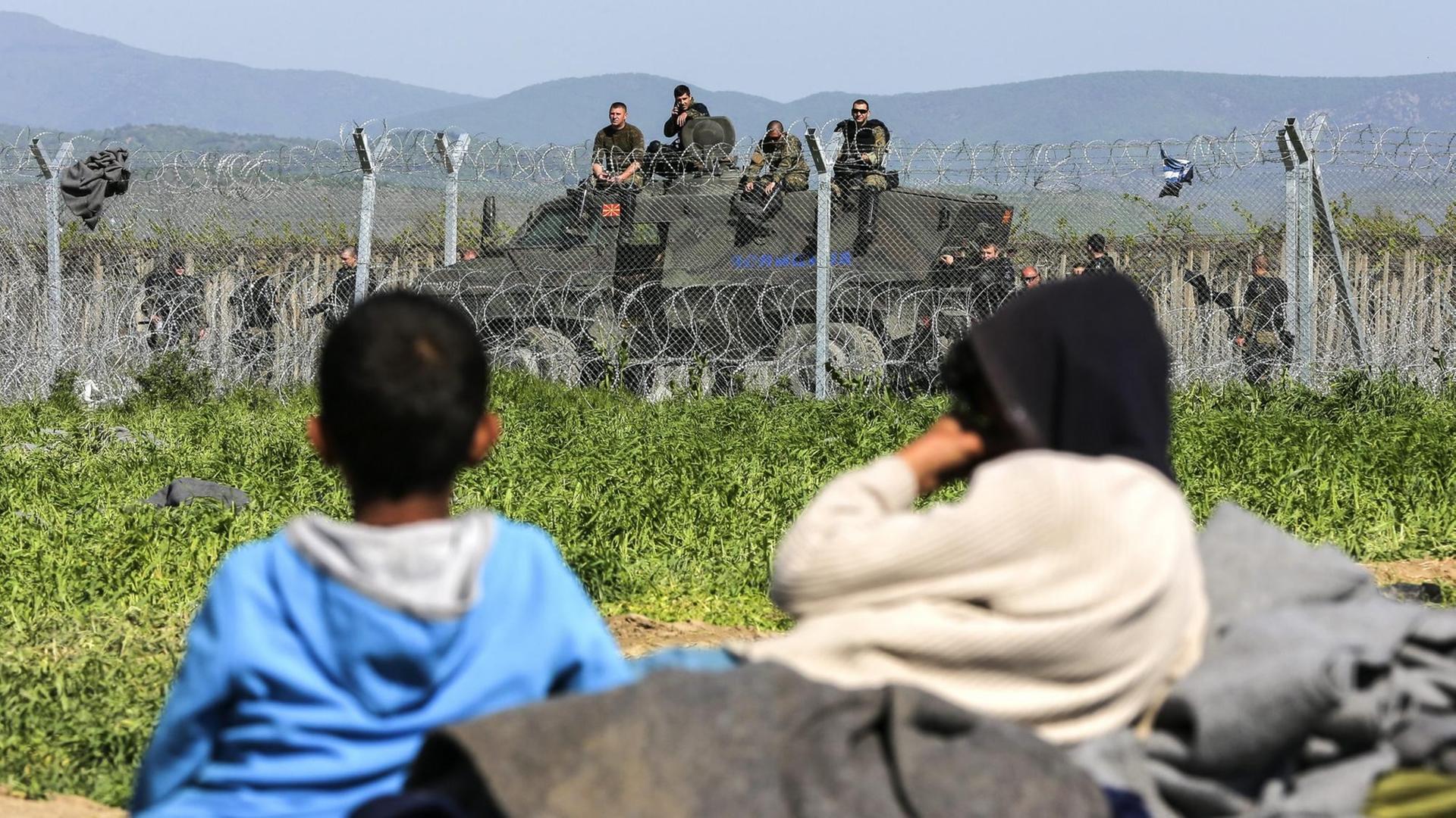 Zwei Flüchtlingskinder liegen im Gras vor einem Stacheldrahtzaun, der Griechenland von Mazedonien trennt. Im Hintergrund ist ein Panzer mit Grenzschutzbeamten zu sehen.