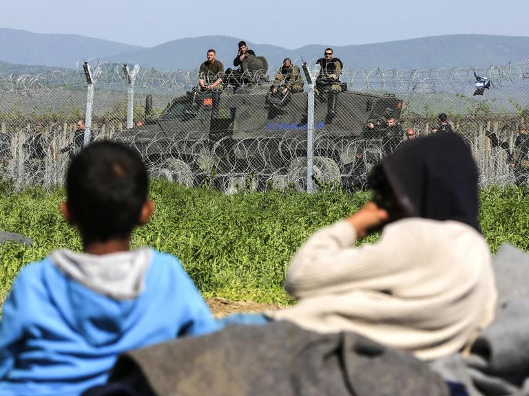 Zwei Flüchtlingskinder liegen im Gras vor einem Stacheldrahtzaun, der Griechenland von Mazedonien trennt. Im Hintergrund ist ein Panzer mit Grenzschutzbeamten zu sehen.