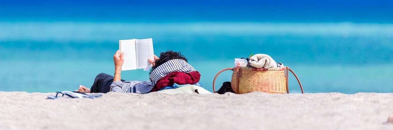 Das Bild zeigt einen Strand. Im Hintergrund ist eine im Sand liegende Frau zu sehen, die ein Buch in der Hand hält und liest. Dahinter ist intensiv blau Himmel und Meer zu sehen.