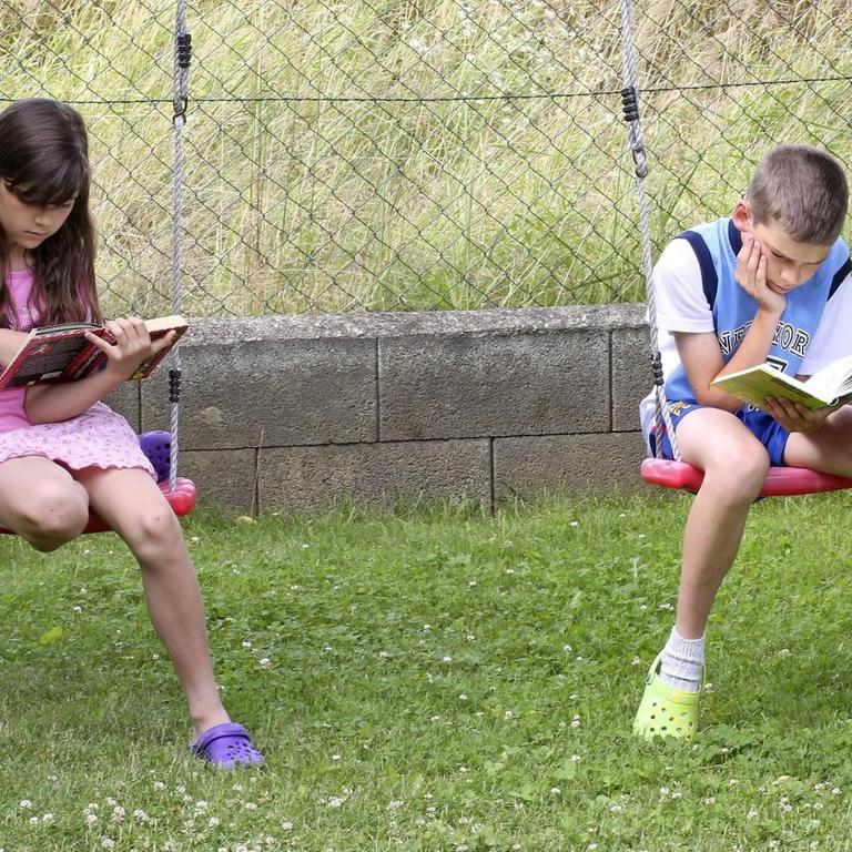 Ein Mädchen und ein Junge lesen Bücher und sitzen dabei auf Schaukeln