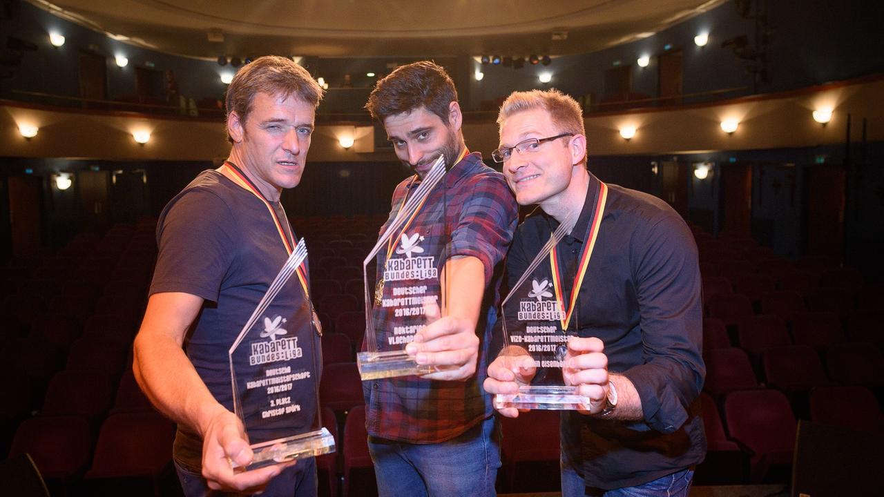 Die Preisträger Christof Spörk, Nektarios Vlachopoulos und Benjamin Eisenberg stehen nebeneinander mit Trophäen der Kabarettbundesliga in der Hand und schneiden Grimassen.