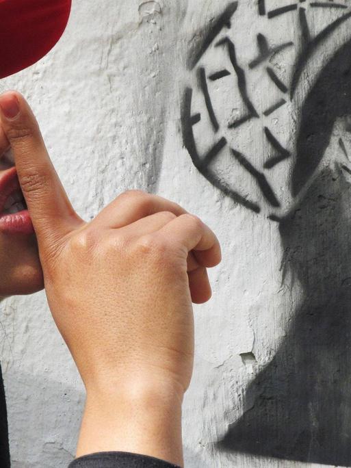Sonita Alizadeh steht im Profil vor einer weißen Wand, auf der ein Graffito gesprüht ist. Sie trägt eine rote Basecap und hält den Zeigefinger ihrer rechten Hand an den Mund. Das Graffito zeigt die gleiche Pose.