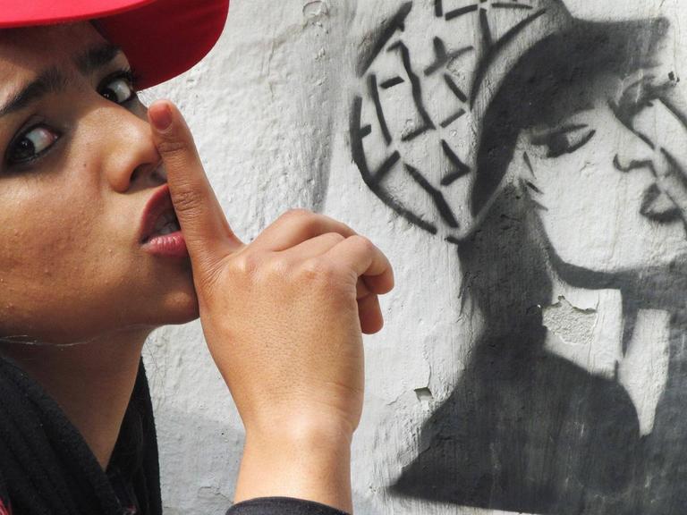 Sonita Alizadeh steht im Profil vor einer weißen Wand, auf der ein Graffito gesprüht ist. Sie trägt eine rote Basecap und hält den Zeigefinger ihrer rechten Hand an den Mund. Das Graffito zeigt die gleiche Pose.