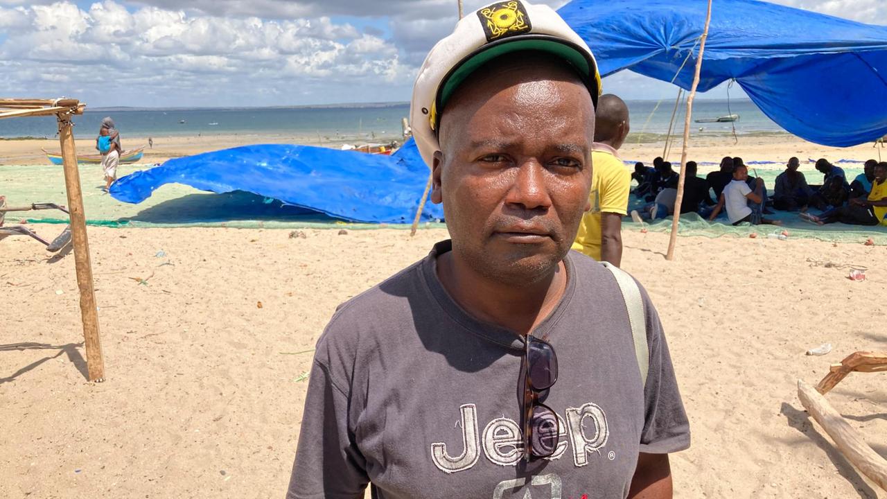 Vasquinho King steht am Strand und schaut ernst in die Kamera. Im Hintergrund sieht man Menschen unter einem blauen Sonnenschutz sitzen.