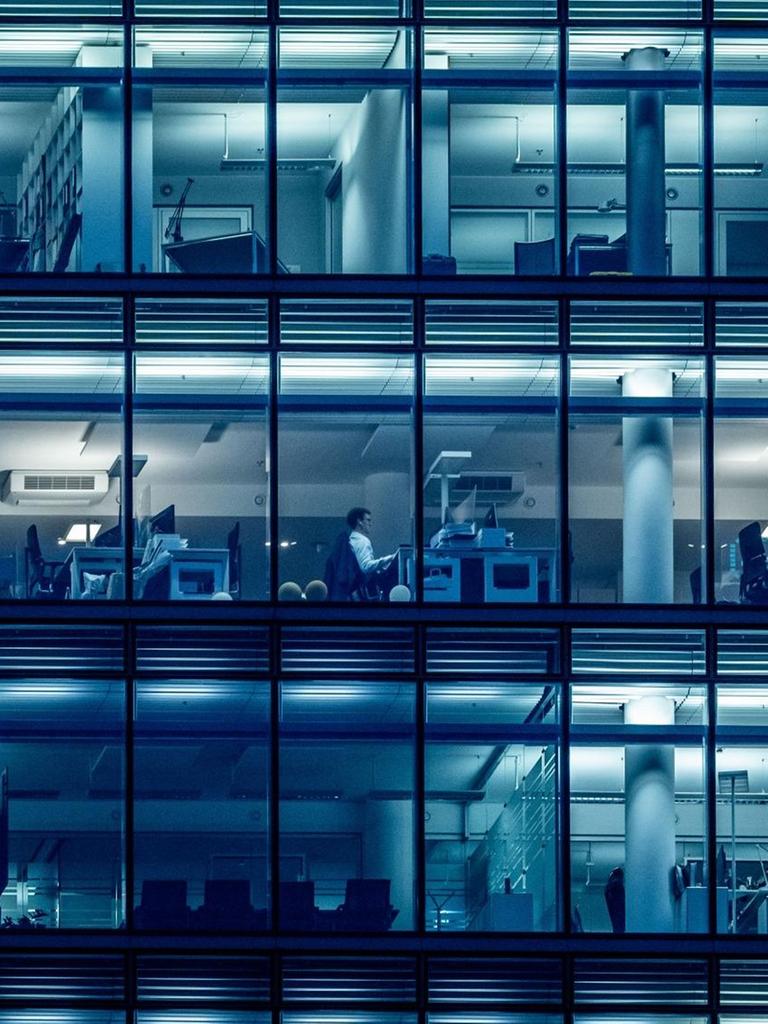 In einem Bürogebäude sind durch die Fenster einzelne Menschen zu sehen, die an Schreibtischen sitzen.