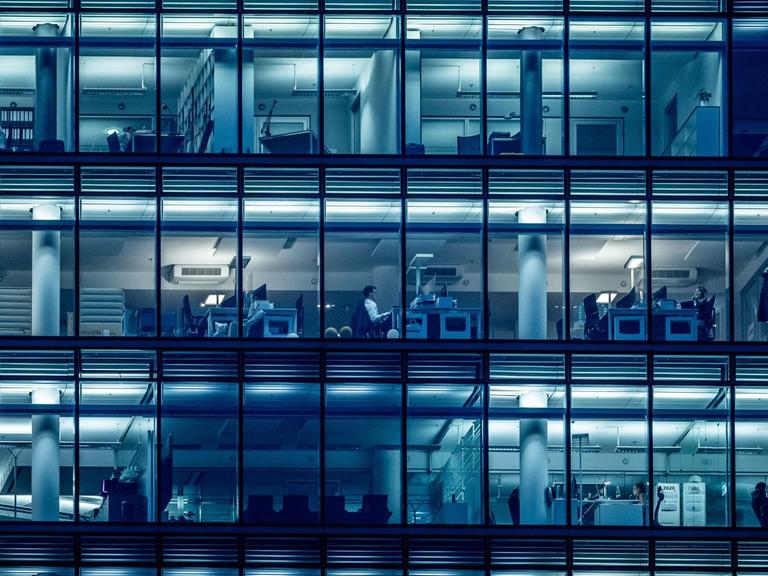 In einem Bürogebäude sind durch die Fenster einzelne Menschen zu sehen, die an Schreibtischen sitzen.