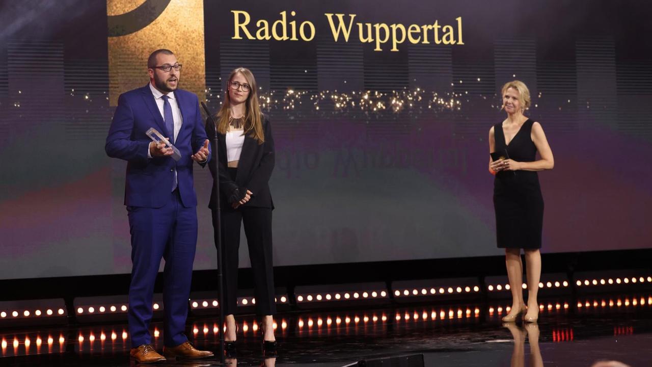 Jens Voss und Laura Mertens von Radio Wuppertal stehen bei der Gala des Deutschen Radiopreises 2021 mit ihrer Auszeichnung an einem Mikrophon.