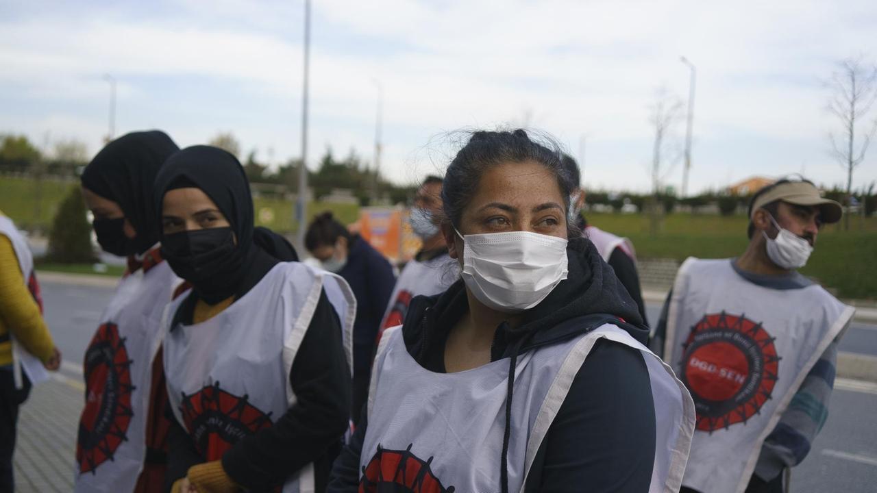 Mehrere Frauen mit Mundschutz und weißen bedruckten Umhängen stehen an einer Straße.