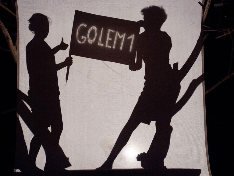 Szene aus "Golem": Eine Frau und ein Mann halten ein Schild mit der Aufschrift "Golem1" - alles ist als Schattenriss zu sehen.