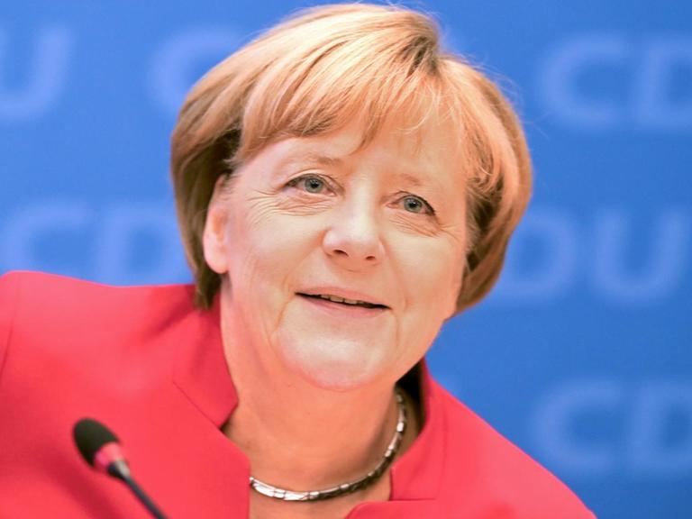 Merkel lächelnd im roten Kostüm vor einem Mikrofon, dahinter eine blaue Wand mit "CDU-Schriftzügen".