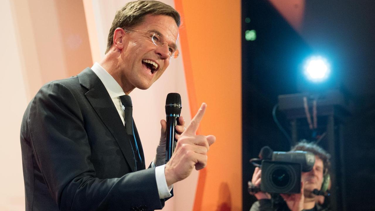 Mark Rutte feiert seinen Wahlsieg vor Anhängern in Den Haag. Er steht auf einer Bühne vor dem organgefarbenen Emblem seiner Partei.