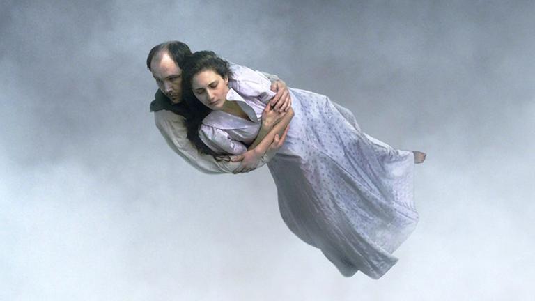 Ein Mann hält eine Frau im langen hellen Kleid im Arm. Beide schweben fast waagerecht in der Luft.
