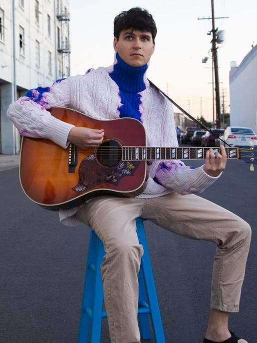 Der Frontmann von Vampire Weekend, Ezra Koenig, spielt mitten auf einer Straße auf einem blauen Hocker Gitarre