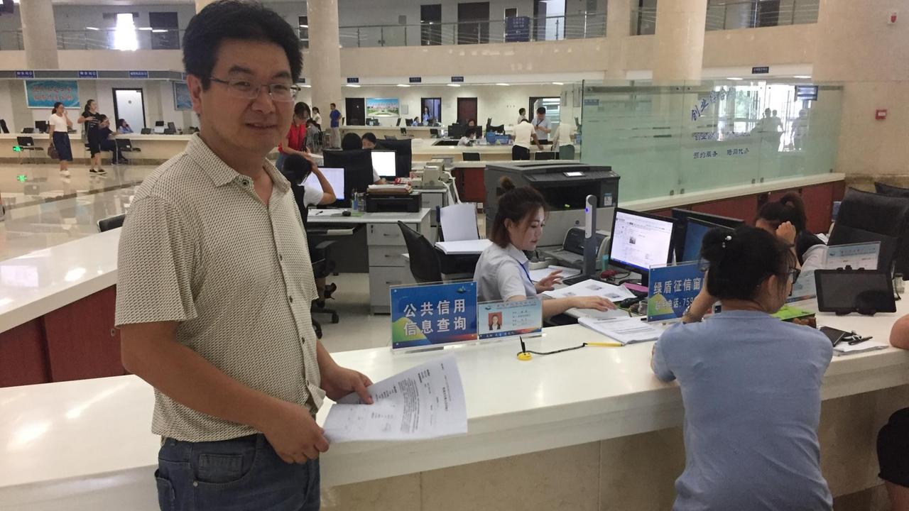 Der Forstamt-Mitarbeiter Zhang Jian trägt ein weißes Hemd und steht im Bürgeramt, in dem viele Frauen an Computern sitzen.