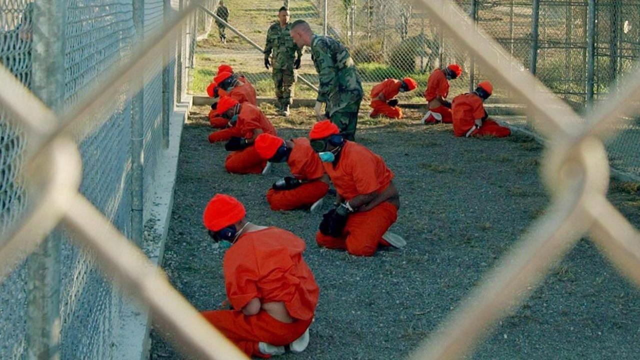 Gefangene im US-Lager Guantanamo auf Kuba knien mit verbundenen Augen hinter Zäunen und Stacheldraht auf dem Boden