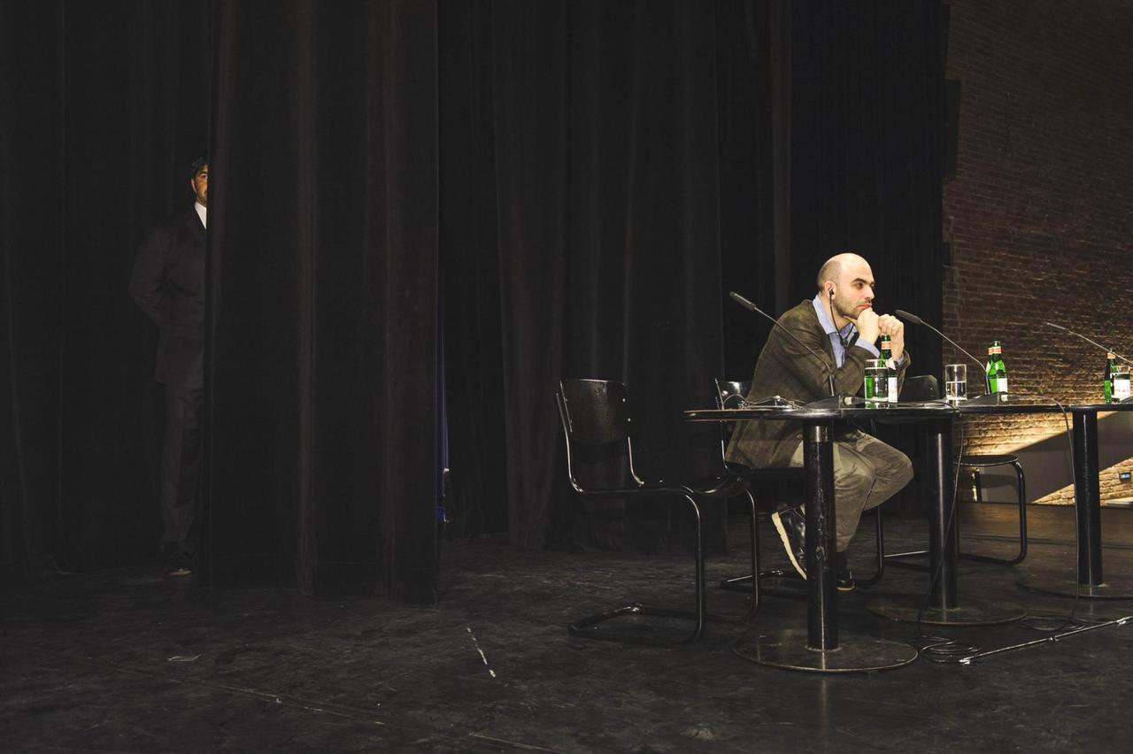 Der italienischen Journalist Roberto Saviano bei einer Lesung im März 2012 in der Akademie der Künste in Berlin. Im Hintergrund steht ein Polizist als Begleitschutz.
