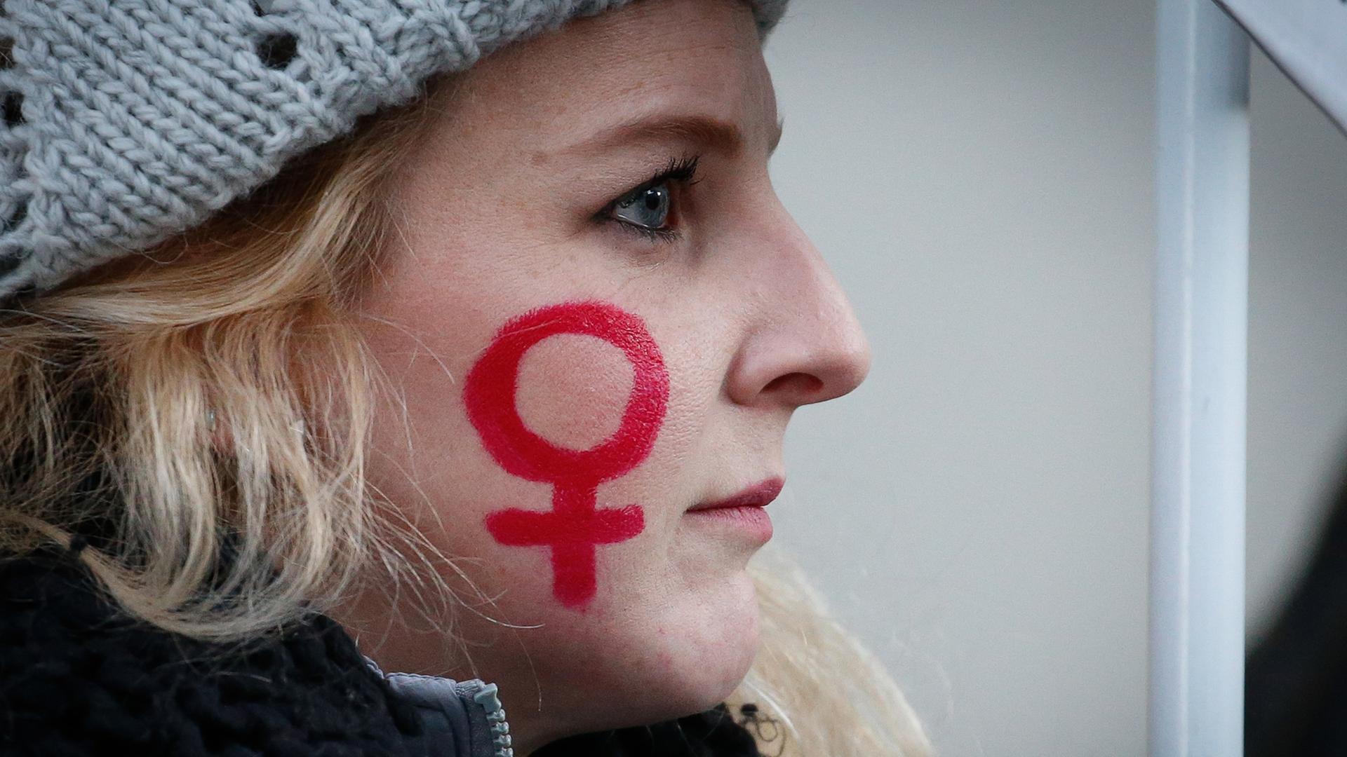 Das Bild zeigt das Foto einer jungen Frau, auf deren Wange mit rotem Stift das weibliche Gender-Symbol gezeichnet ist.
