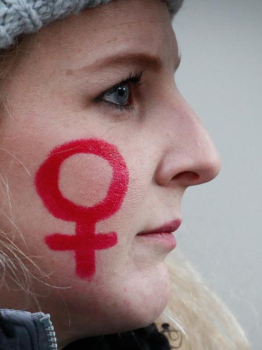Das Bild zeigt das Foto einer jungen Frau, auf deren Wange mit rotem Stift das weibliche Gender-Symbol gezeichnet ist.