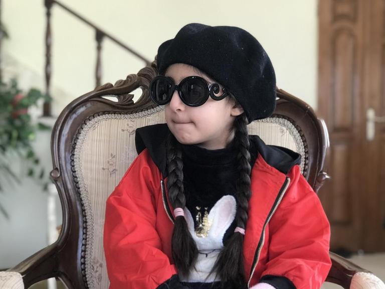 Ein kleines Mädchen in roter Jacke, mit dunkler Brille und schwarzem Käppi sitzt auf einem Stuhl.