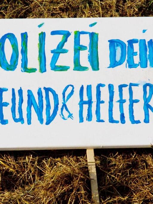 "Polizei dein Freund und Helfer?", steht geschrieben auf einem Schild in blauen Buchstaben und mit rotem Fragenzeichen auf einer grünen Wiese liegend.