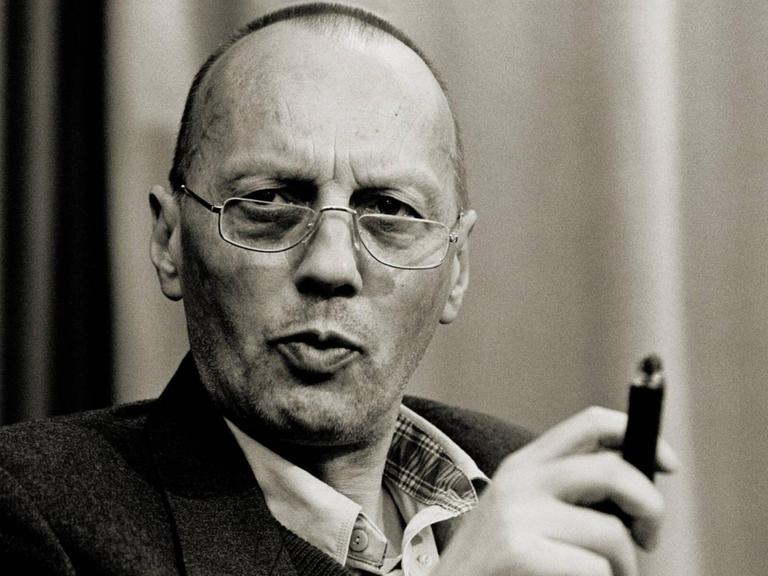 Schwarzweißporträt von Karl Mickel, der mit ernster Miene und einer Zigarre in der Hand in die Kamera blickt.