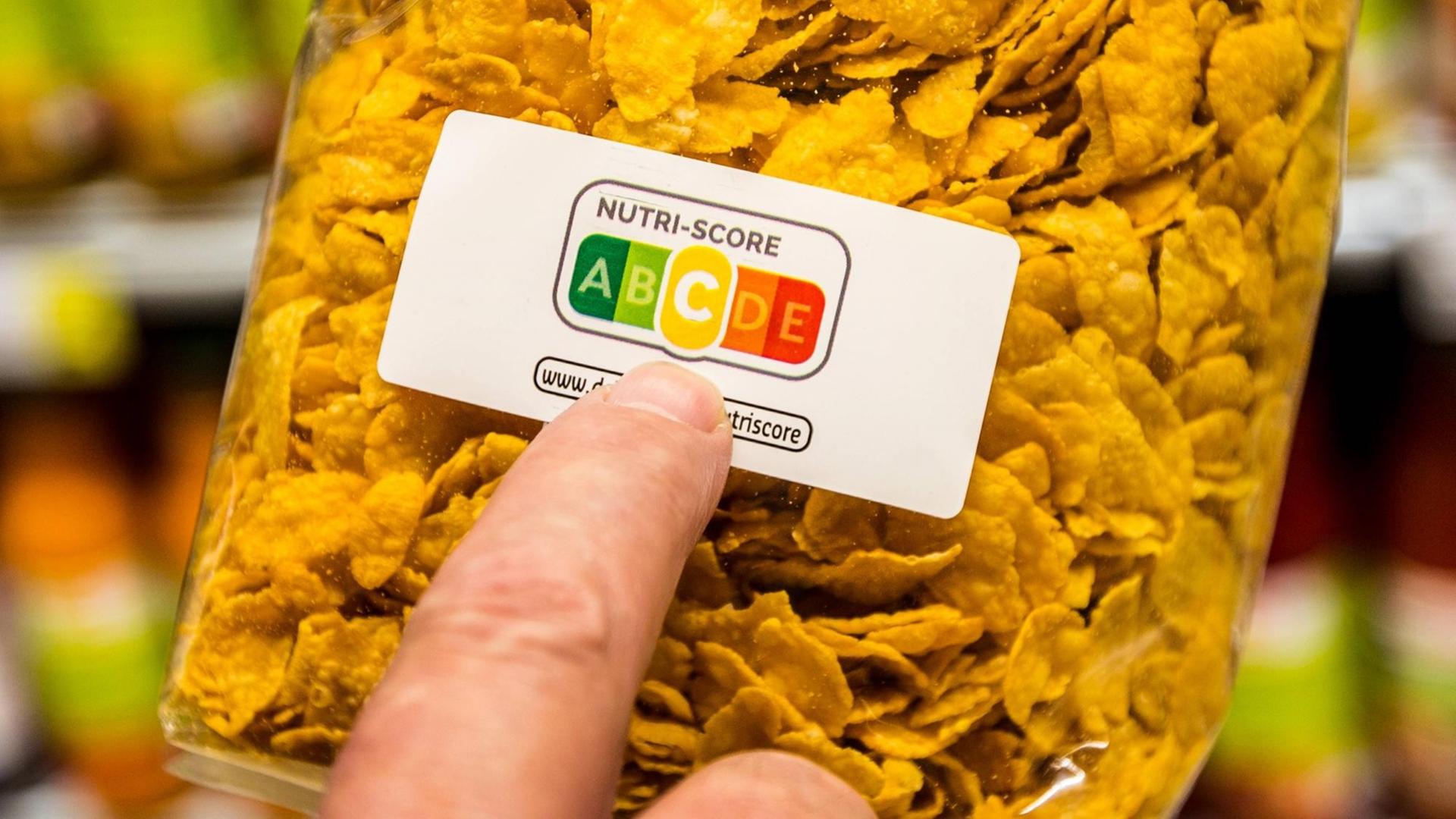 Eine Packung Cornflakes, ein Zeigefinger deutet auf die Farbflächen des Nutri-Score-Aufdrucks. Die mittlere Kategorie "C" (gelb) ist hervorgehoben.