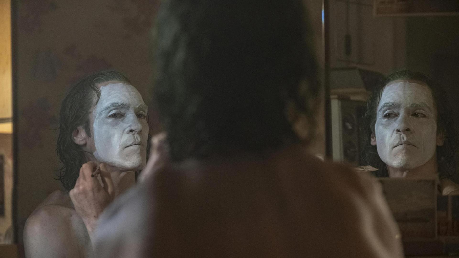 Schauspieler Joaquin Phoenix schminkt sich als Joker vor einem Spiegel. Man sieht ihn von hinten und zweimal von vorne als Spiegelbild.