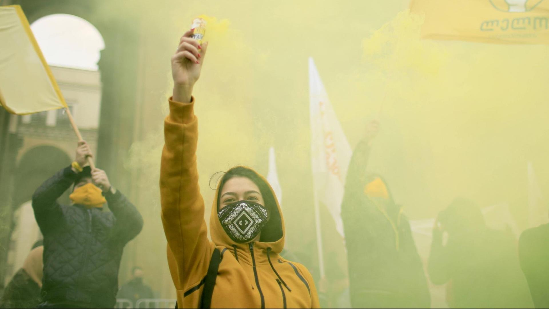 Eine junge Frau in gelber Jacke reckt bei einer Demonstration den Arm in die Höhe.
