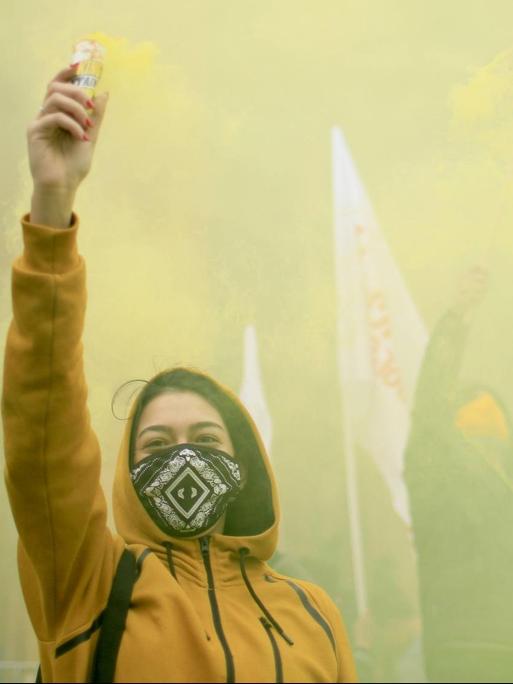 Eine junge Frau in gelber Jacke reckt bei einer Demonstration den Arm in die Höhe.