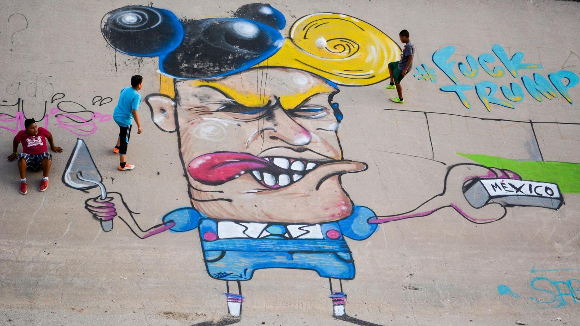 Kinder spielen am 23.06.2016 neben einer Graffiti-Karikatur des designierten US-Präsidenten Donald Trump im Flussbett des Rio Grande (Rio Bravo in Mexiko). Ein Streetart-Künstler sprayte die neun Meter große Karikatur an die Wand des zubetonierten Kanals an der Grenze zwischen Mexiko und den USA in der Stadt Ciudad Juarez.
