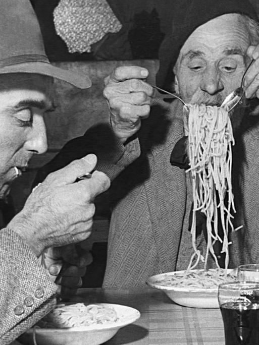 Eine historische Fotografie von zwei Männern, die an einem Tisch Spagetti essen.