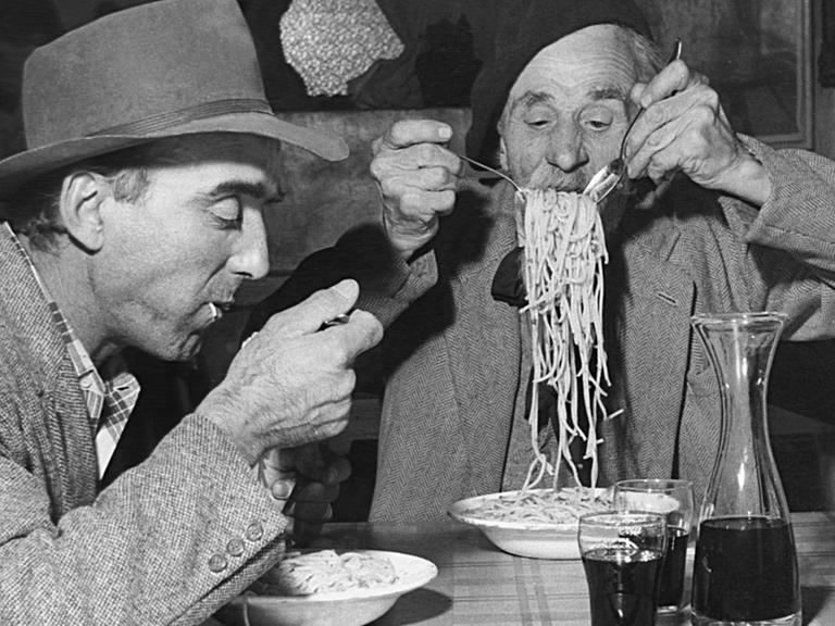 Eine historische Fotografie von zwei Männern, die an einem Tisch Spagetti essen.